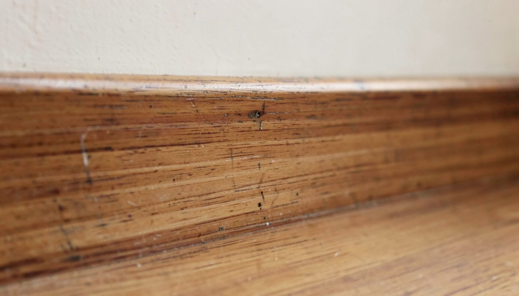 A wooden baseboard with subtle surface damage and faint mud-like tracing, warm brown and tan tones, quiet interior setting.